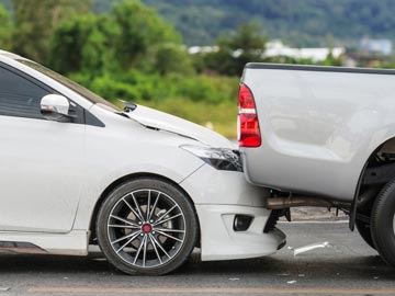 Ein weißer Sedan wird von einem silbernen Pickup auf einer Straße aufgefahren, wobei der vordere Teil des Sedans sichtbare Schäden aufweist.